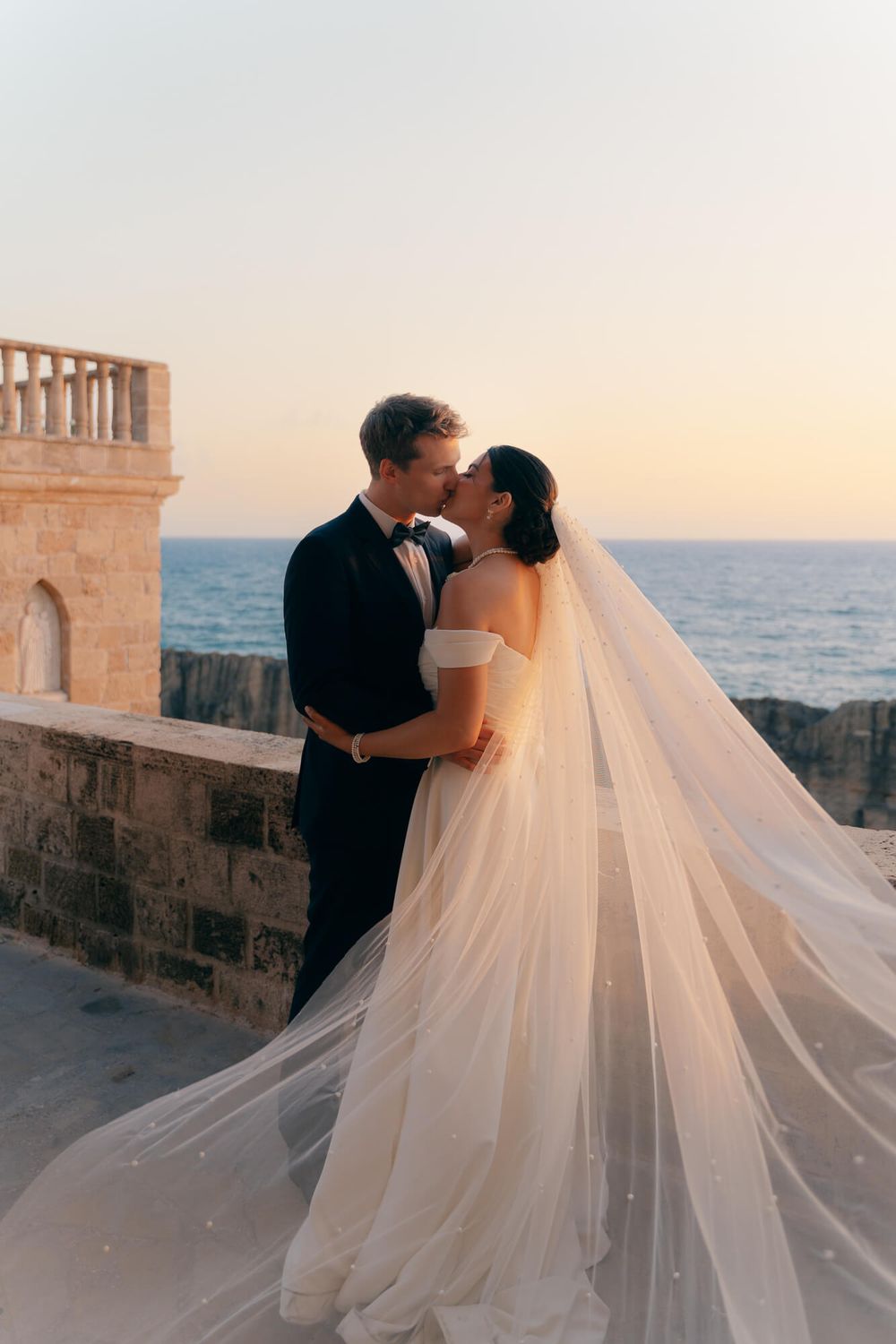 Bride and groom share a romantic kiss at sunset by the ocean, her flowing veil billowing in the breeze.