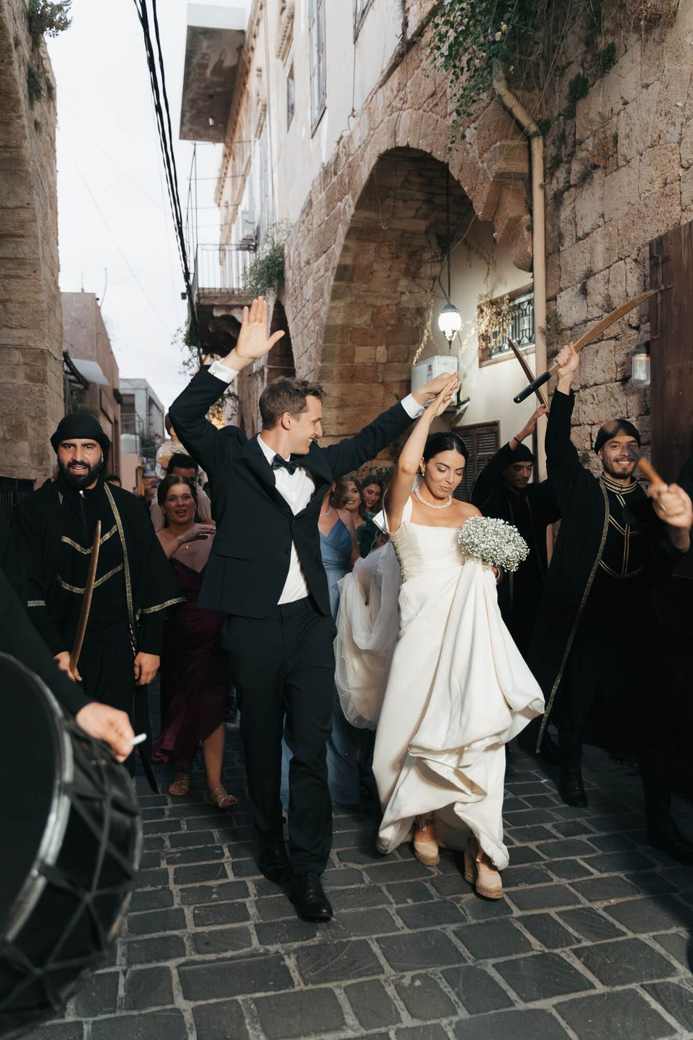 Bride and groom celebrate wedding exit on cobblestone street with musicians in black uniforms and guests cheering.