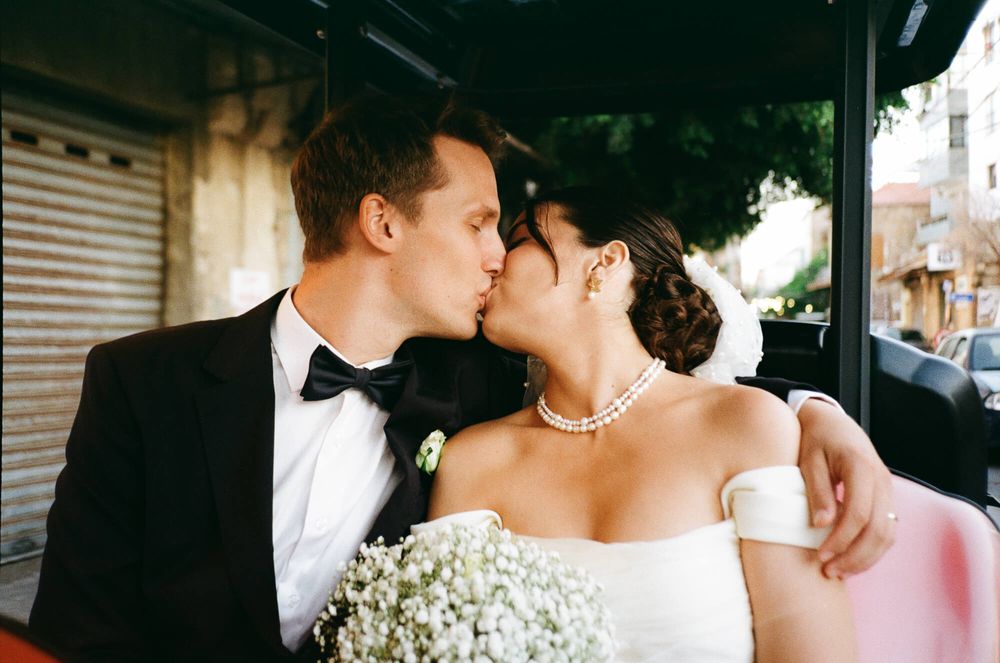 Bride and groom sharing a romantic kiss, bride holding white baby's breath bouquet, wearing pearl necklace and off-shoulder gown.