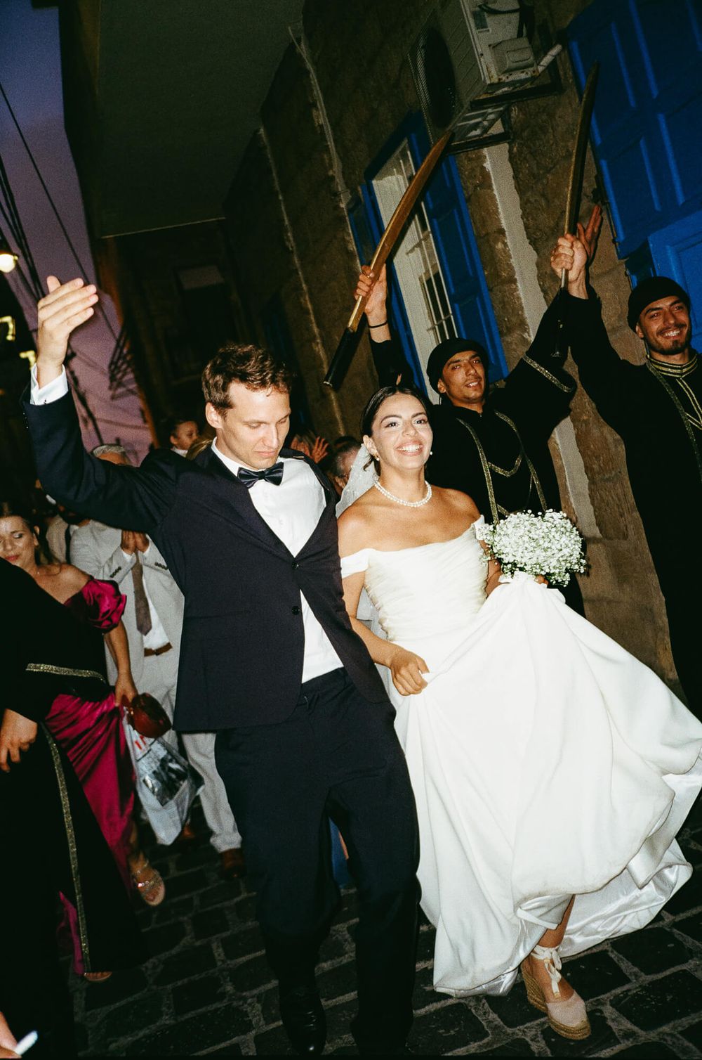 Joyful newlyweds celebrate outdoors at night, bride in white gown holding bouquet, groom in tuxedo, traditional musicians behind.