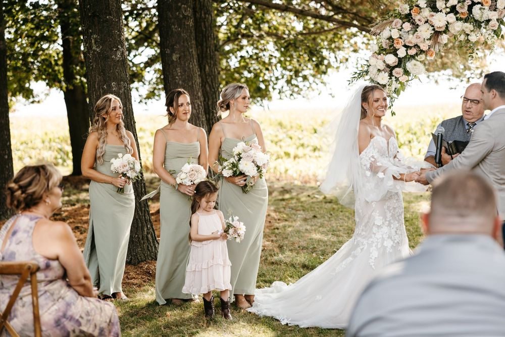 An outdoor wedding ceremony under trees with bridesmaids in sage green dresses and a flower girl in white.