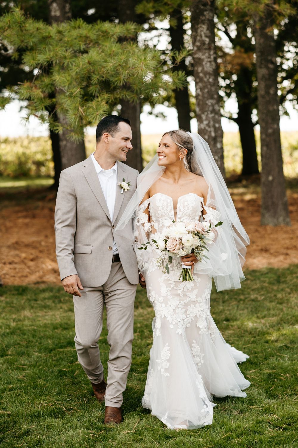 A bride in a lace mermaid gown and groom in light gray suit share a joyful moment outdoors among pine trees.