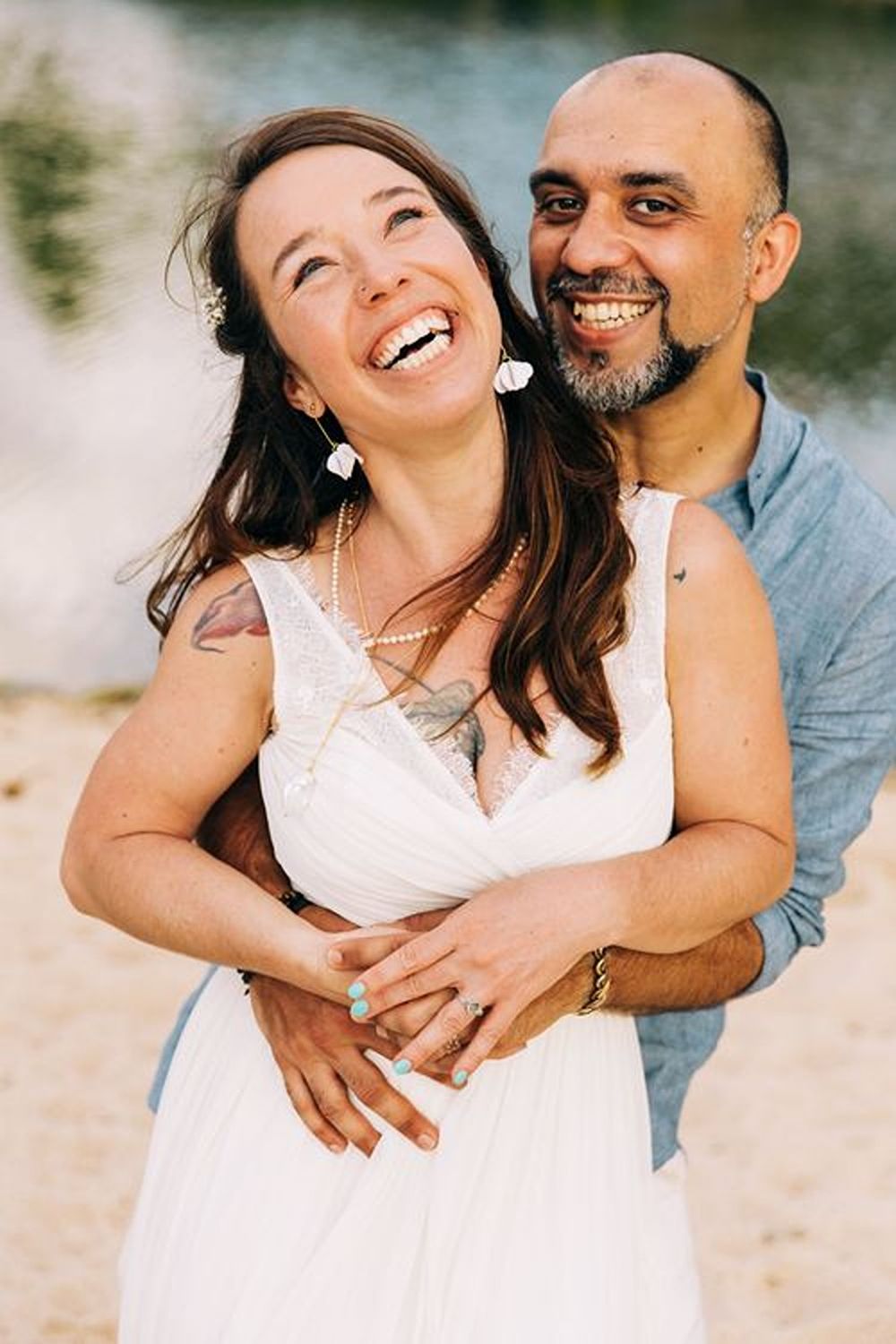 A romantic couple embrace and laugh together on a beach, with the woman wearing a white dress and the man in denim.