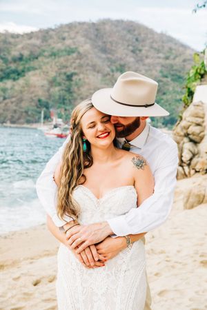 A joyful couple embraces on a tropical beach with mountains and boats in the background.
