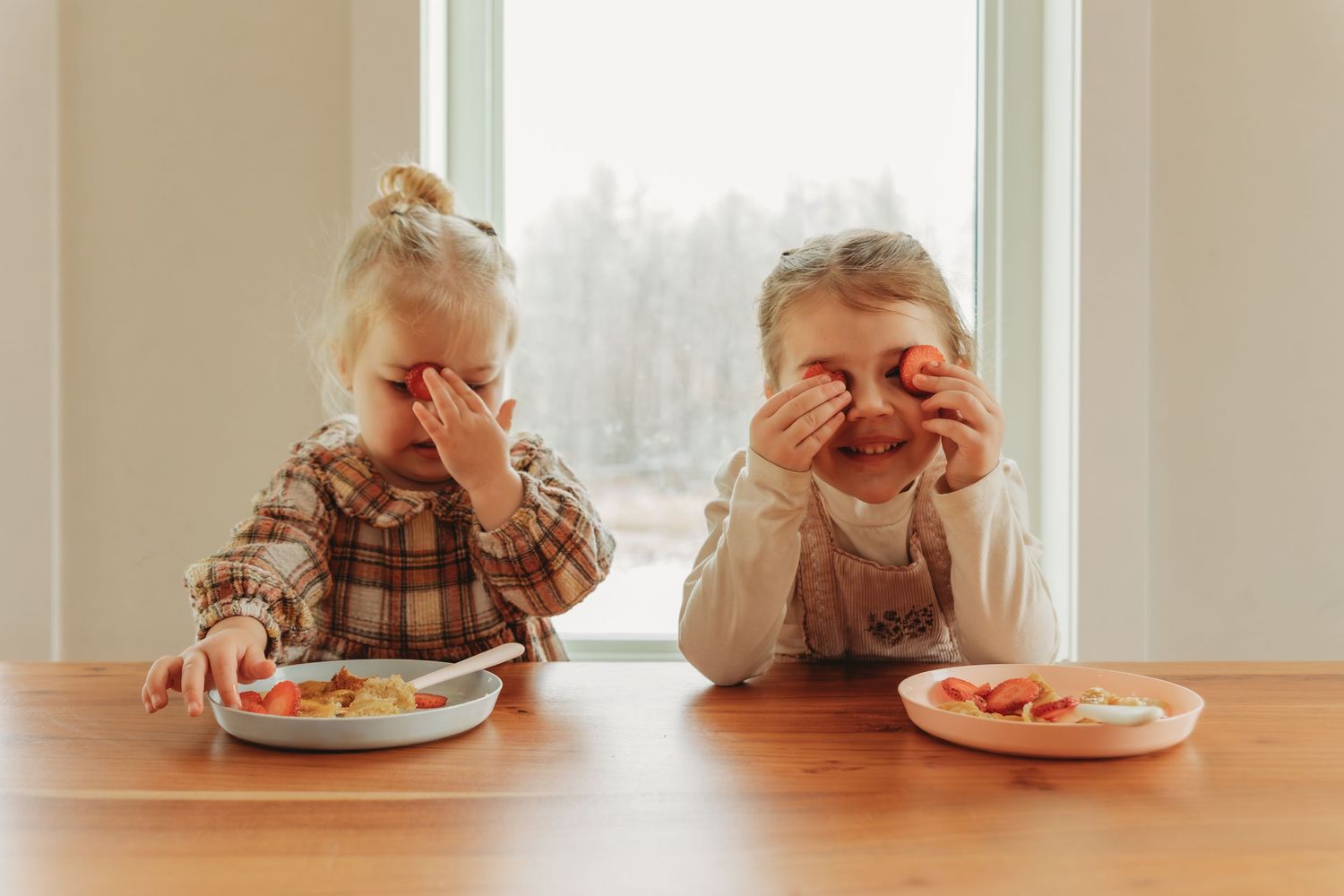 Two young children eating breakfast while making silly faces at a wooden table.