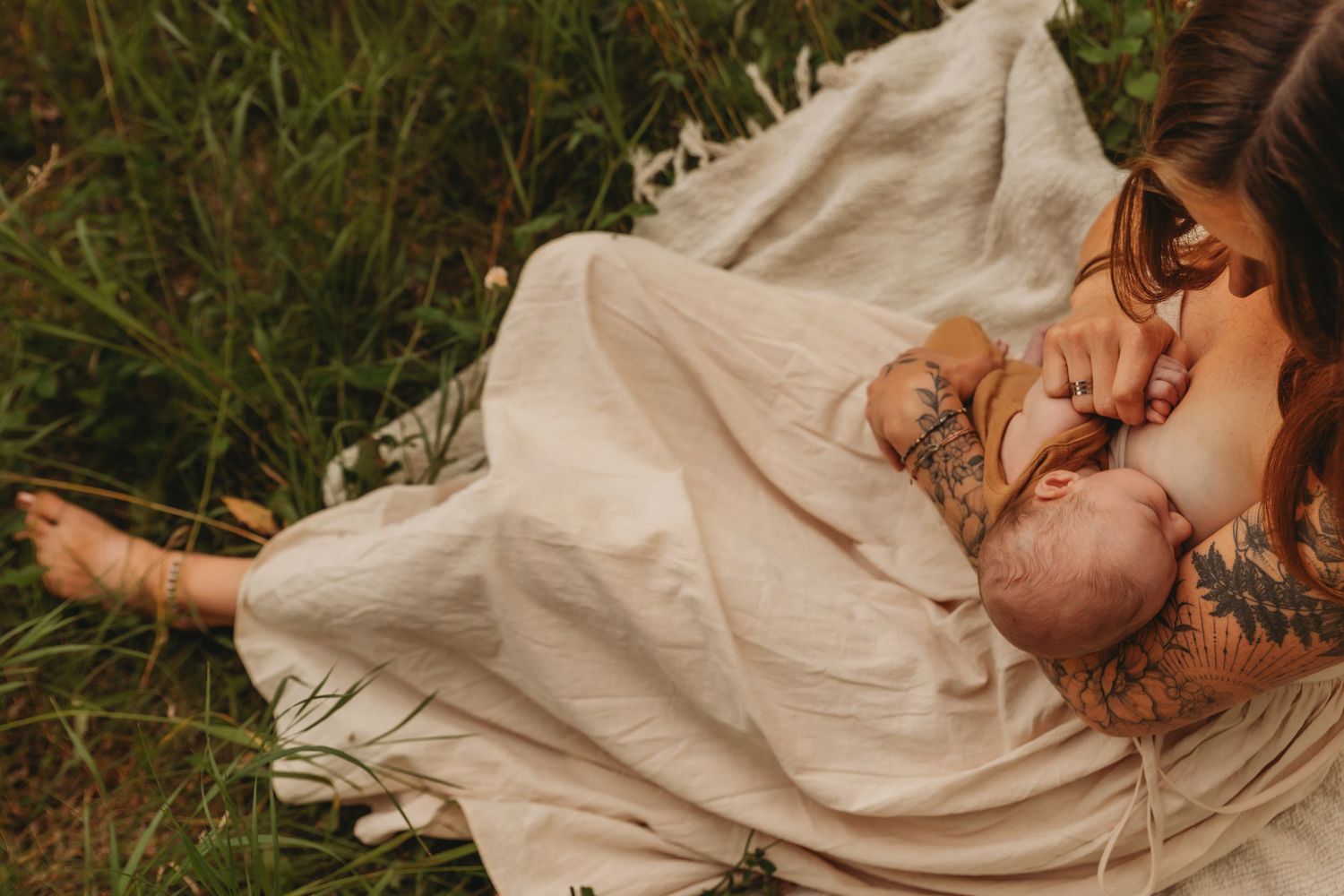 Person in flowing dress resting on grass with newborn baby in natural outdoor setting.
