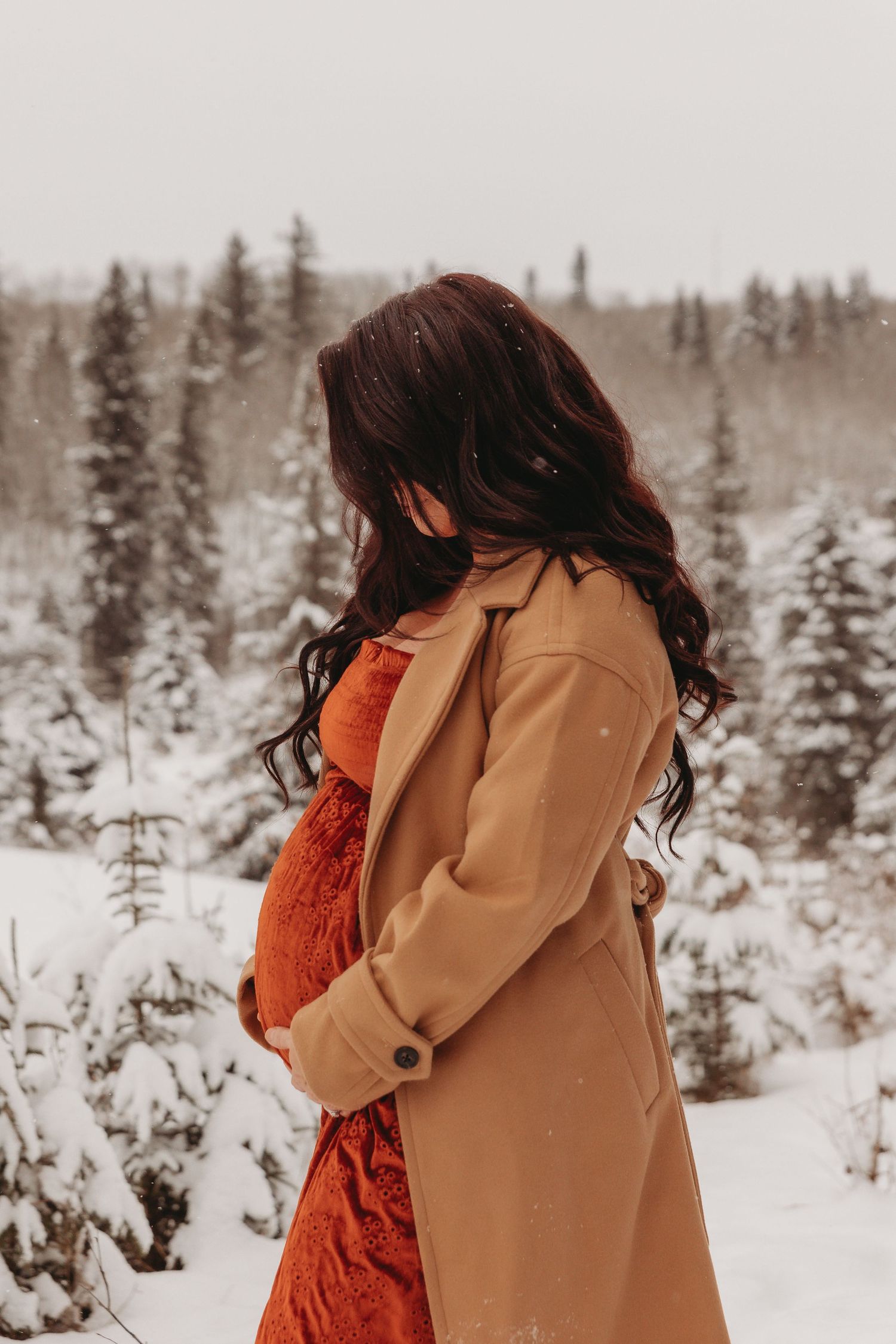 Side view of person in red dress and camel coat standing in snowy winter forest.
