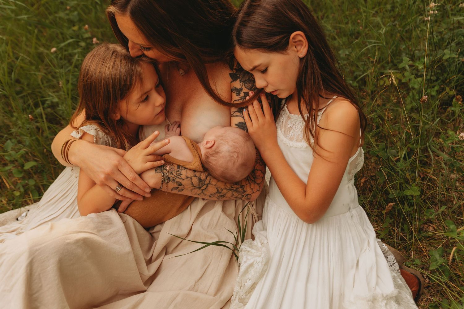Family gathered outdoors in white dresses sharing intimate moment with baby on blanket.