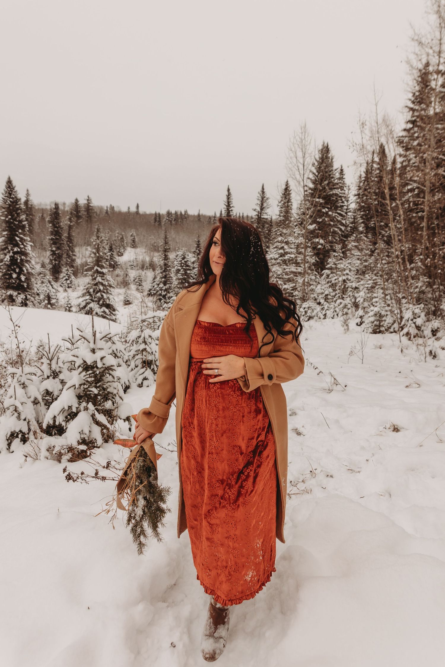Person in red velvet dress and tan coat poses in snowy winter forest landscape.