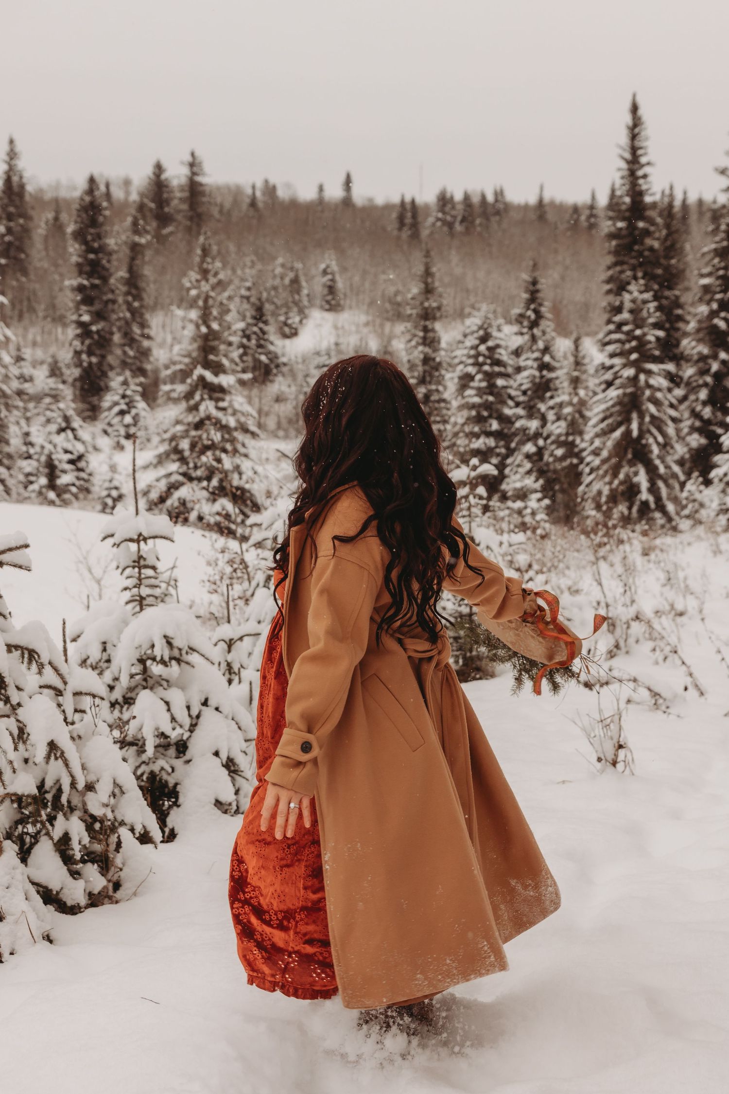 A woman in a tan coat and red velvet dress stands in a snowy forest with evergreen trees in the background.