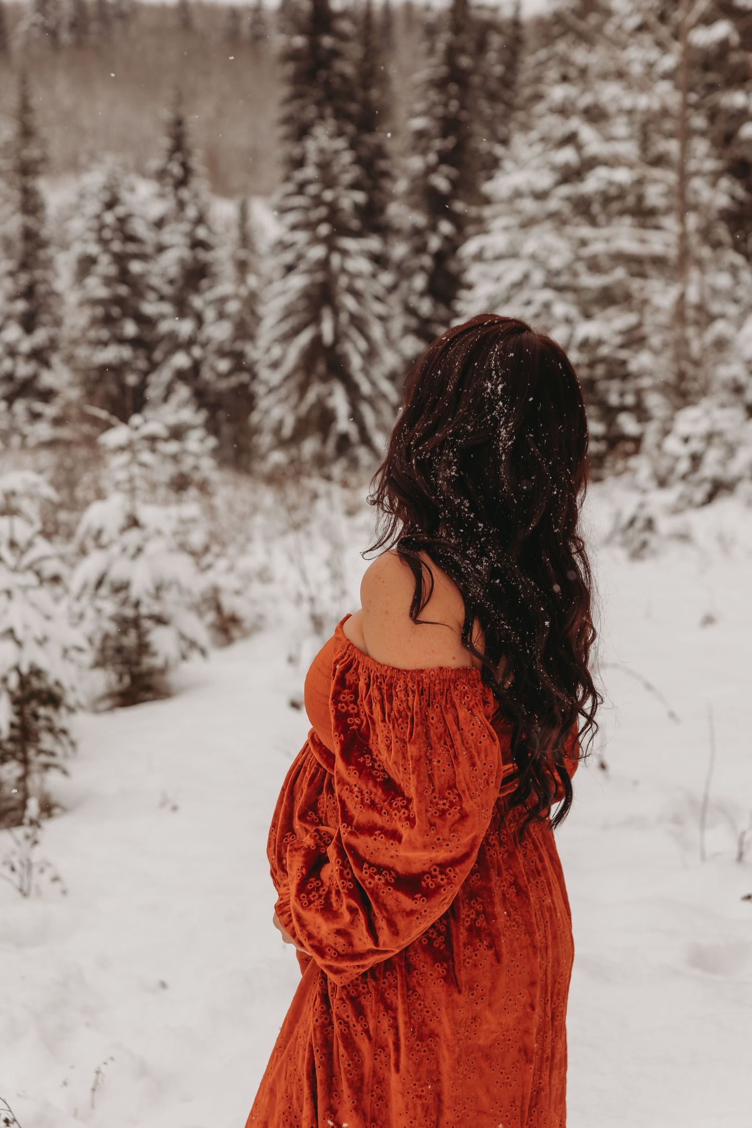 Maternity in a red velvet dress stands in a snowy forest landscape with evergreen trees.