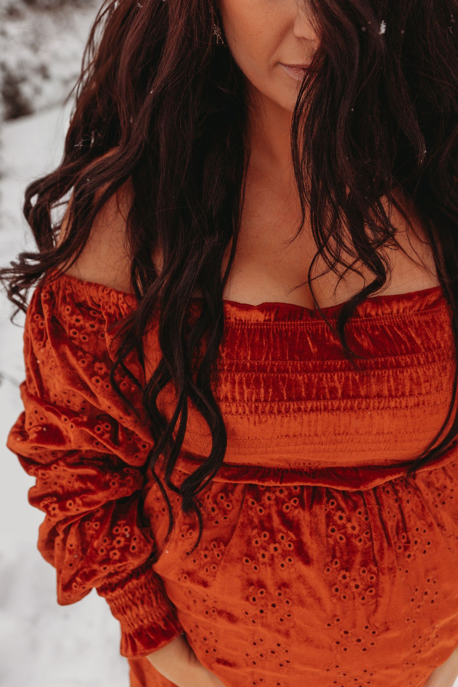 Long dark hair cascades over a red velvet off-shoulder dress against a snowy winter backdrop.