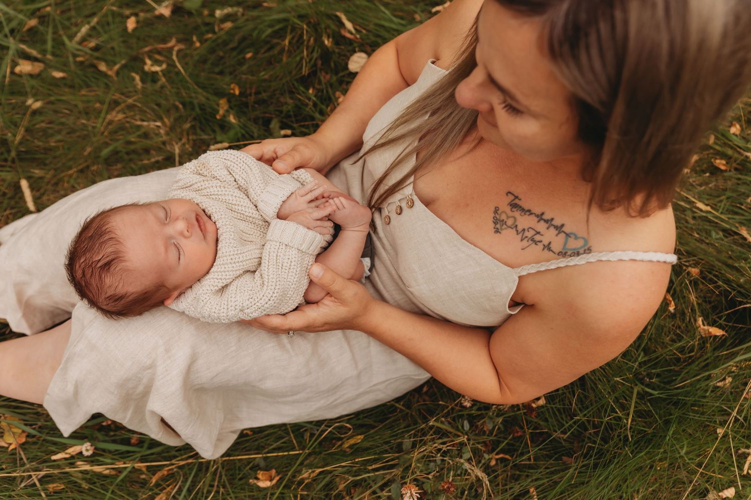 Newborn baby in white knit outfit being cradled on white blanket in grassy outdoor setting.
