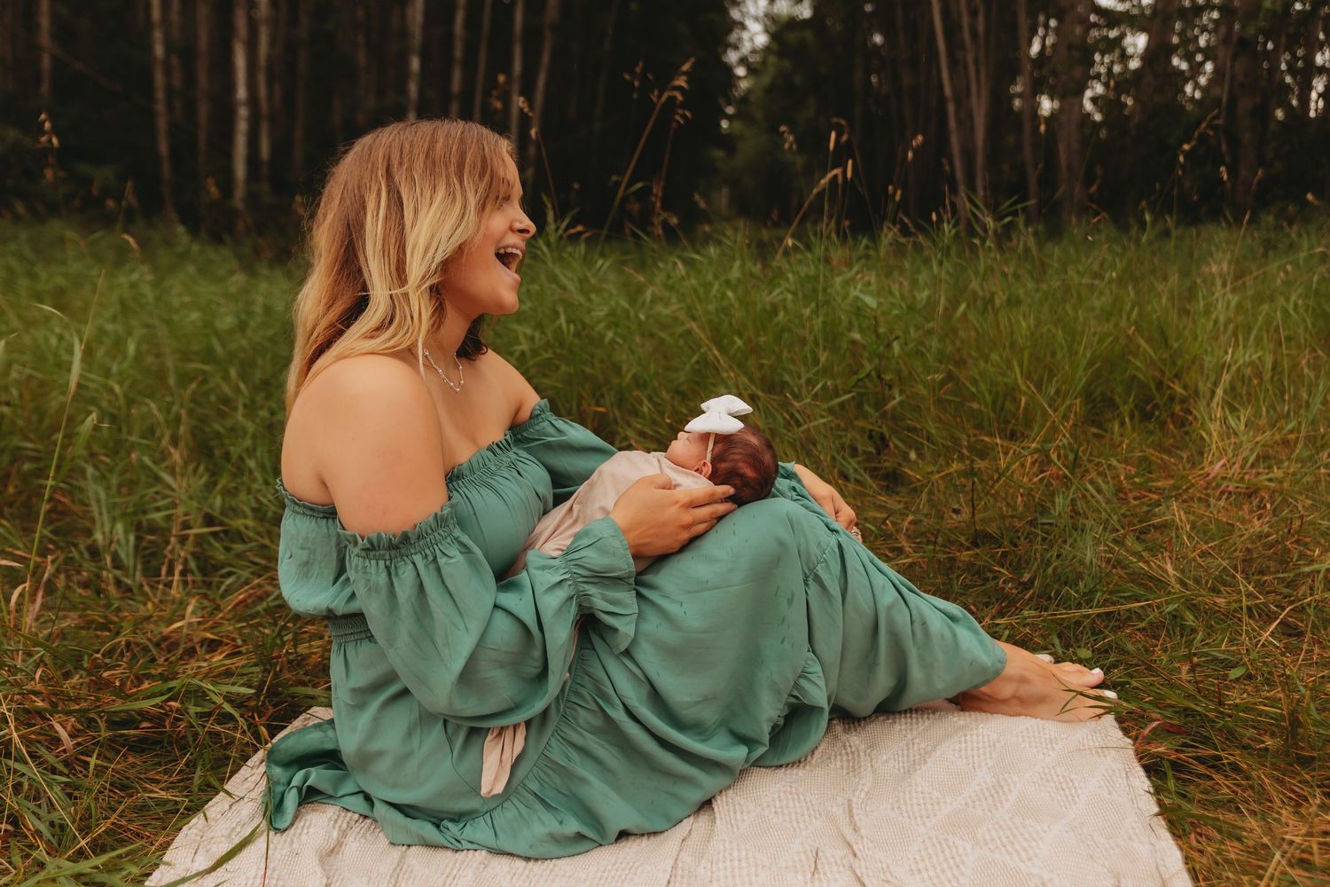 Person in turquoise dress holding baby while sitting on blanket in sunlit forest setting.
