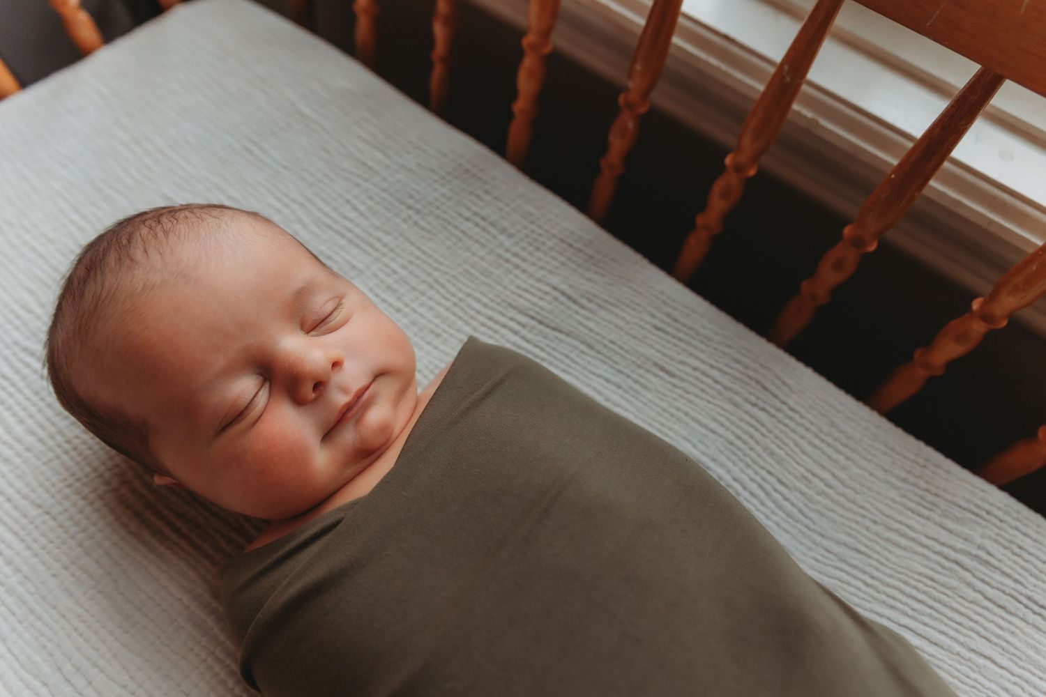 Peaceful sleeping baby wrapped in olive green swaddle lying in wooden crib with plush white bedding.