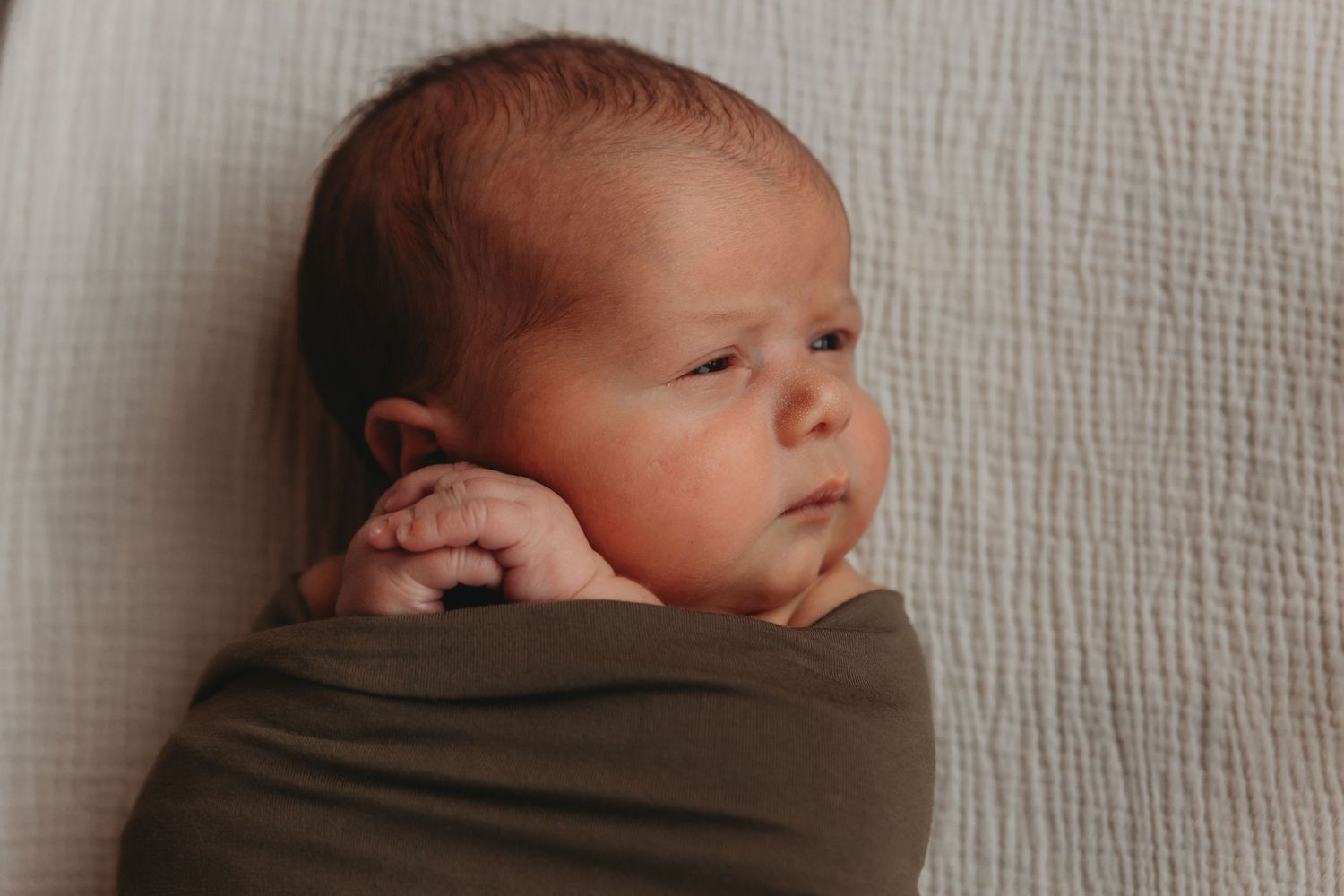 Newborn wrapped in brown swaddle blanket resting on white textured surface.