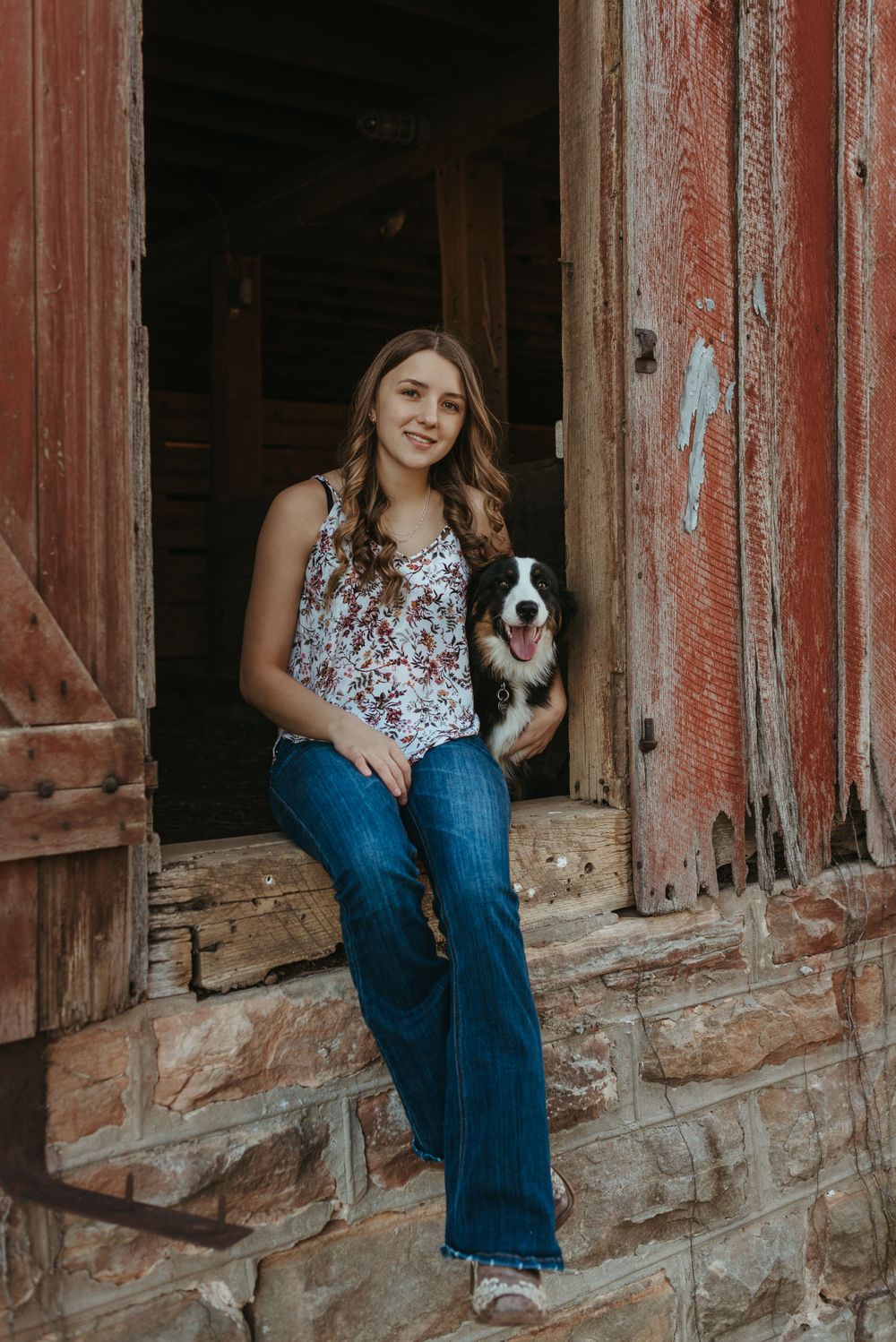A person sits with their spotted black and white dog in front of a rustic red barn doorway.