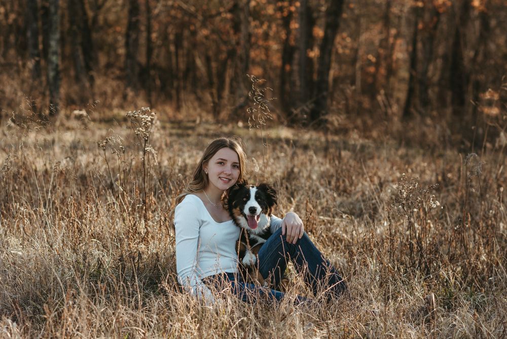 A person and their dog sit together in tall autumn grass at sunset.