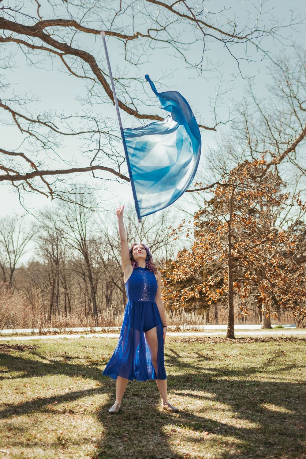 Dancer in flowing blue dress tosses fabric while performing outdoors under bare tree branches.