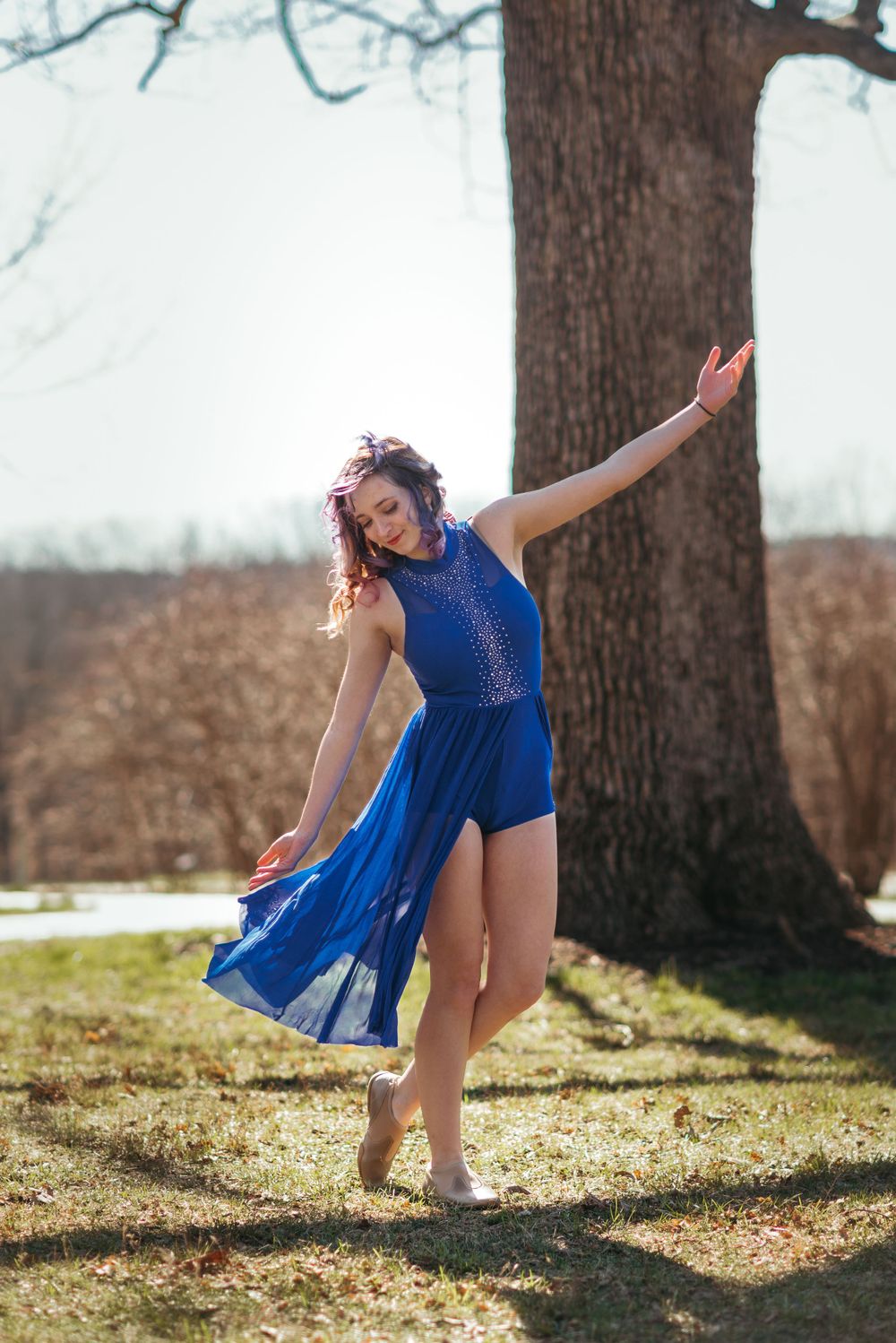 Graceful dancer in royal blue dress poses elegantly near a large tree trunk in natural lighting.
