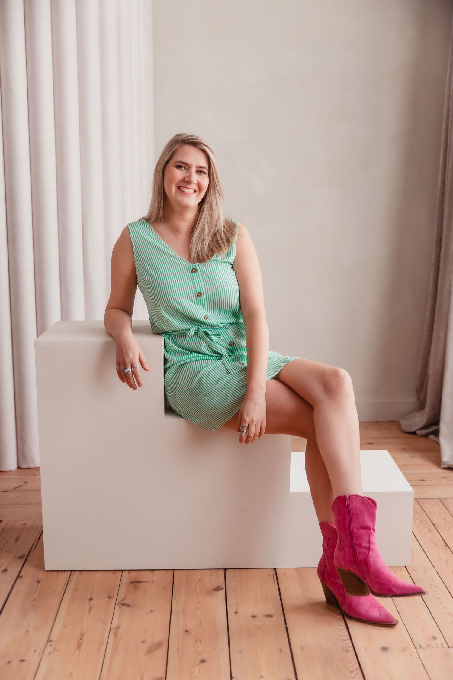 A woman in a mint dress and pink cowboy boots sits on a white cube against a curtained wall.