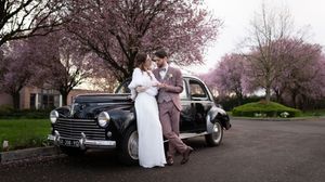 Bride and groom posing beside a vintage black car under blooming pink cherry trees.