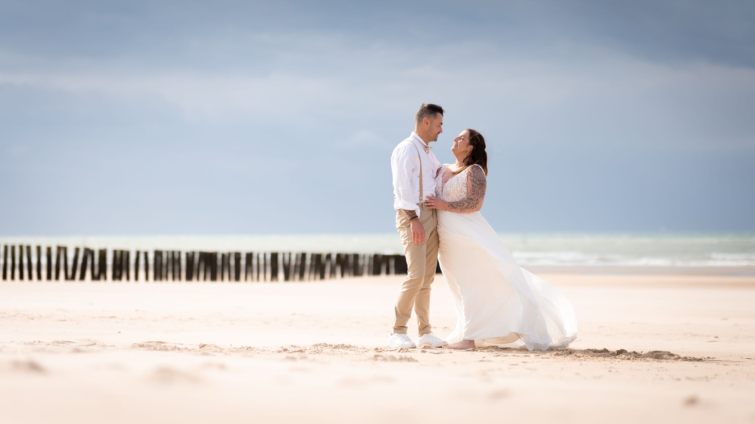 Wedding couple embracing on a sandy beach with wooden posts and cloudy sky backdrop.