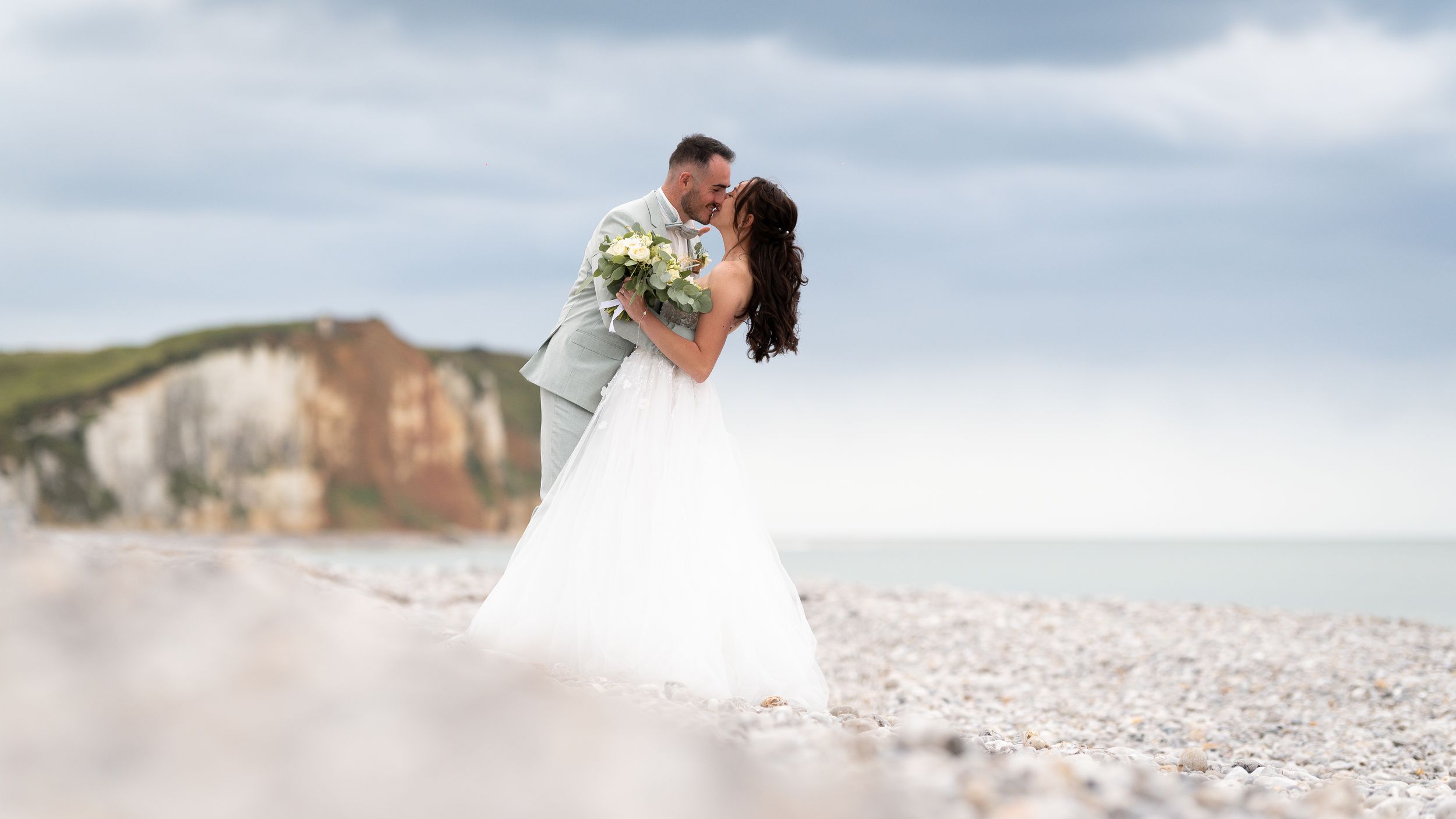 Bride and groom kissing on pebble beach with white chalk cliffs backdrop.