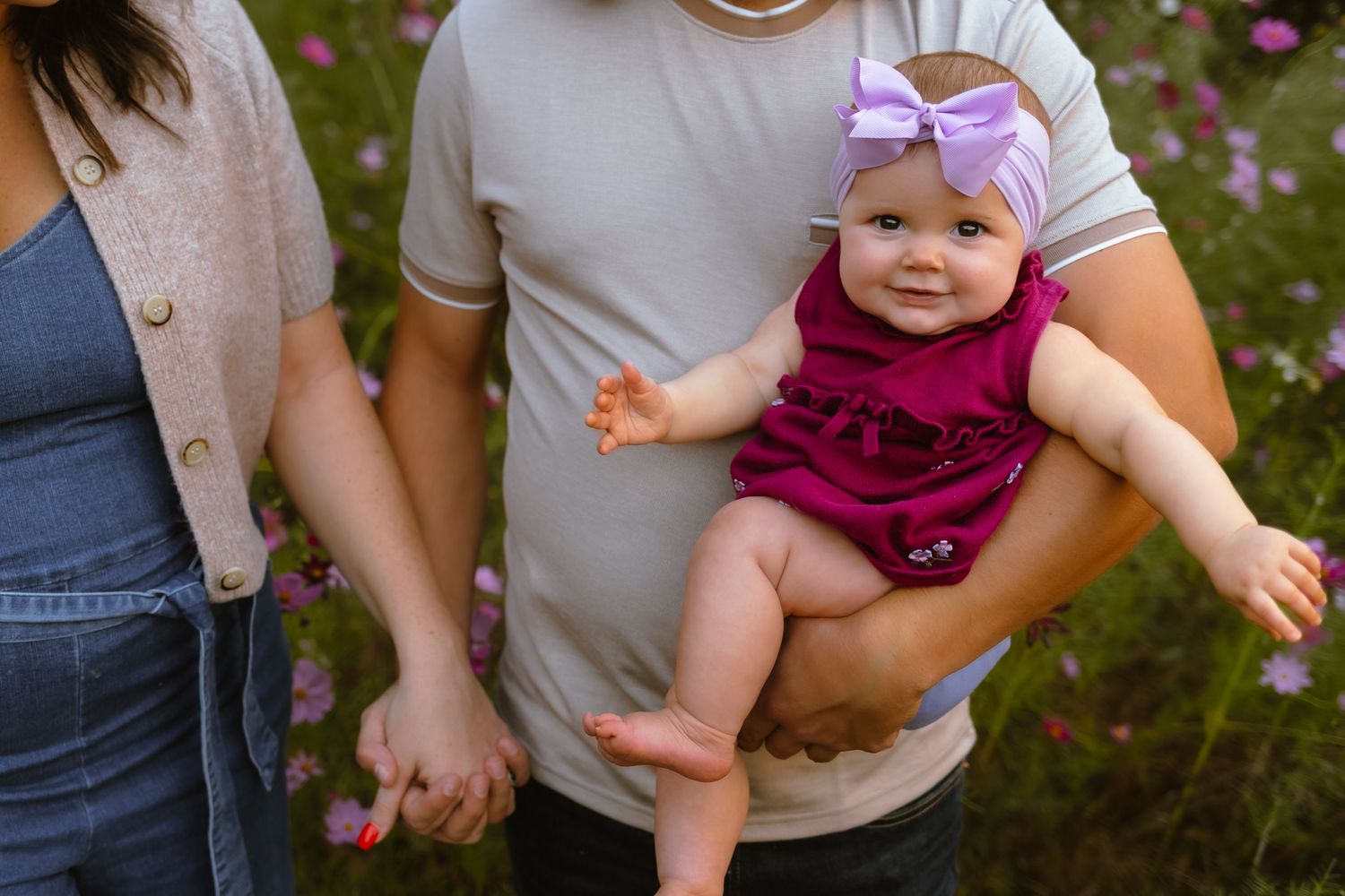 A small baby in a pink dress and bow headband explores pink wildflowers in a garden setting.