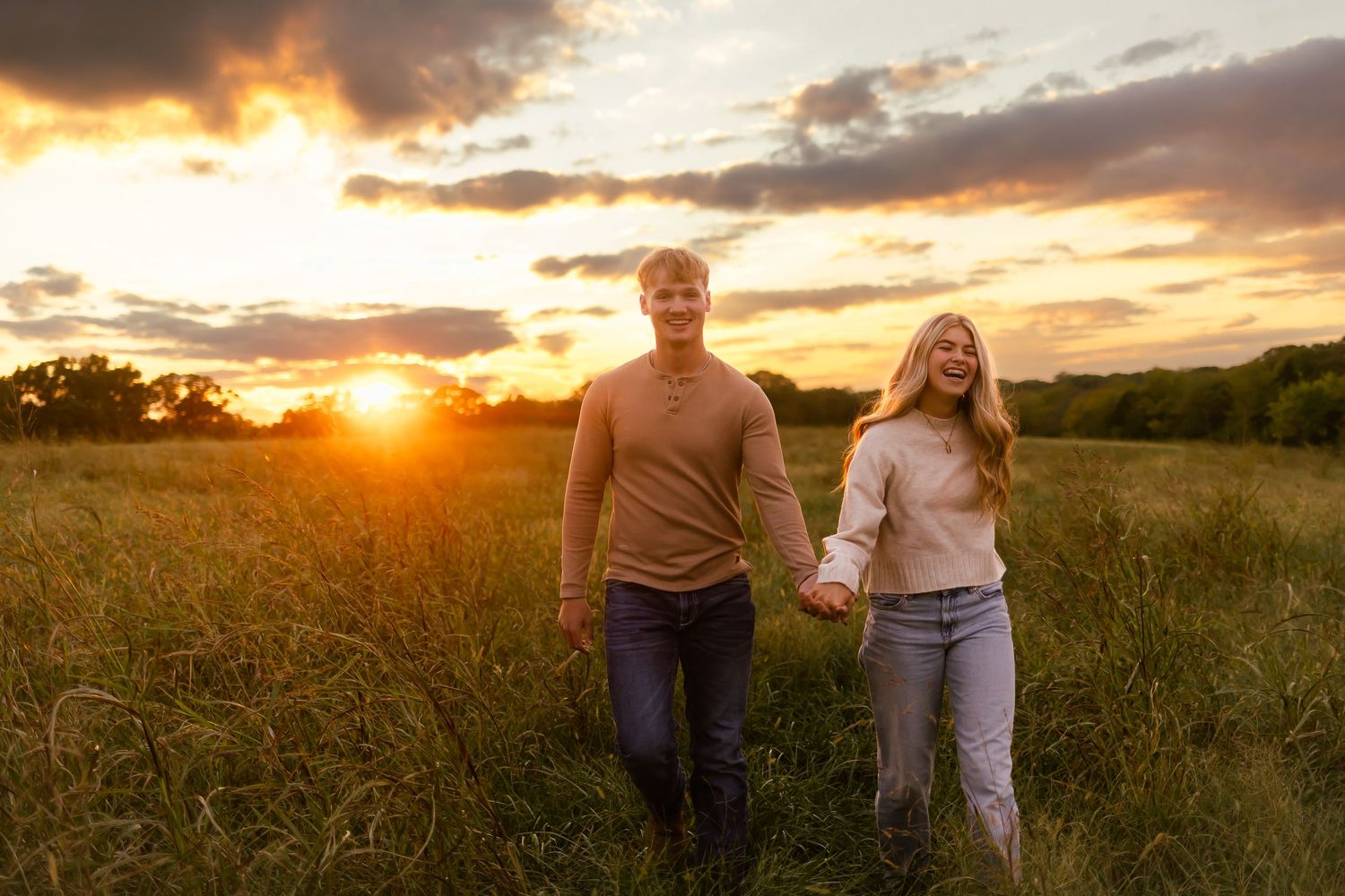 A couple walks hand in hand through a golden field at sunset with dramatic clouds in the sky.