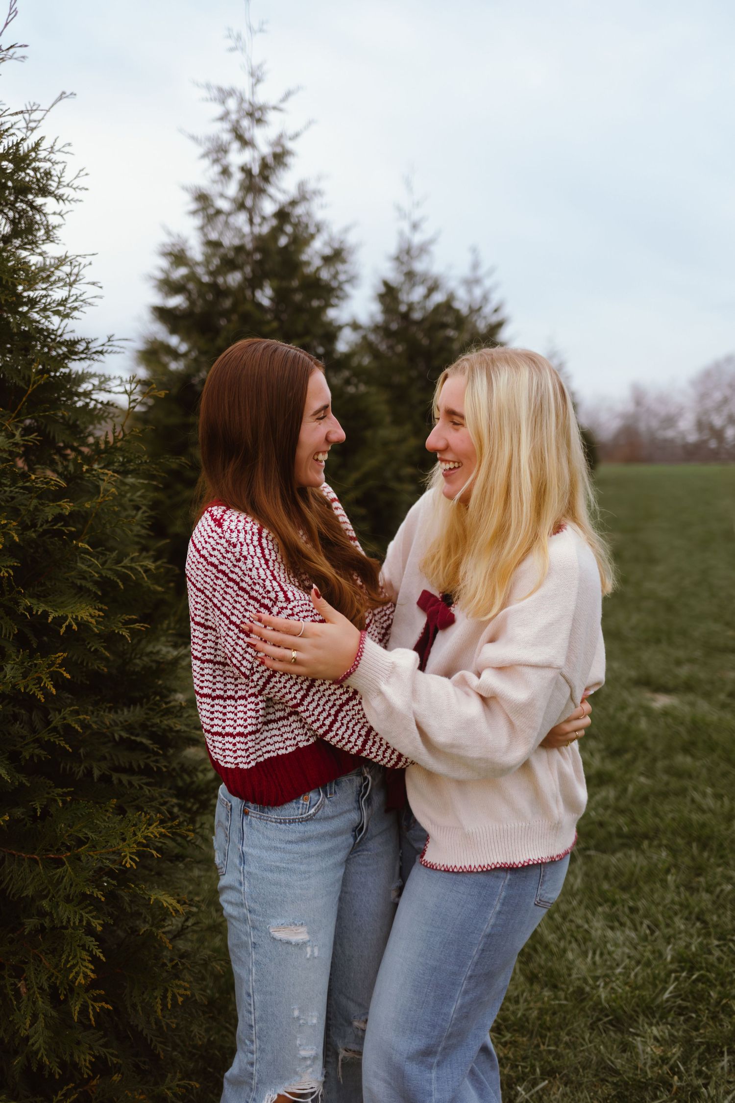 Two friends in casual outfits share a joyful moment while standing near evergreen trees in a grassy field.