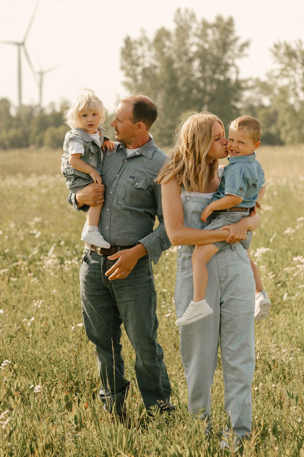 Family in casual denim outfits standing together in wildflower field with wind turbine visible in background.