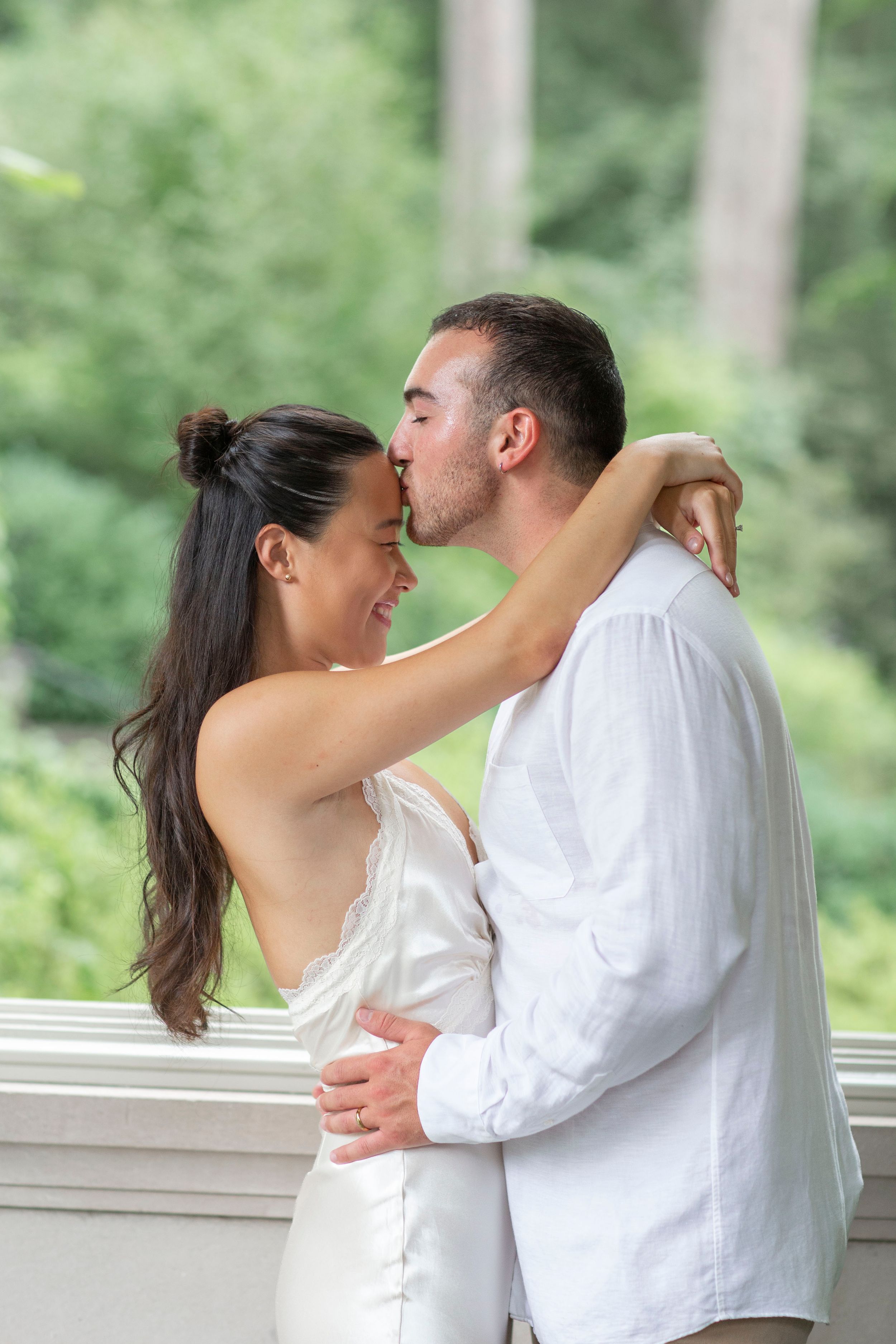 Groom kisses smiling bride on the forehead while embracing outdoors in green surroundings.