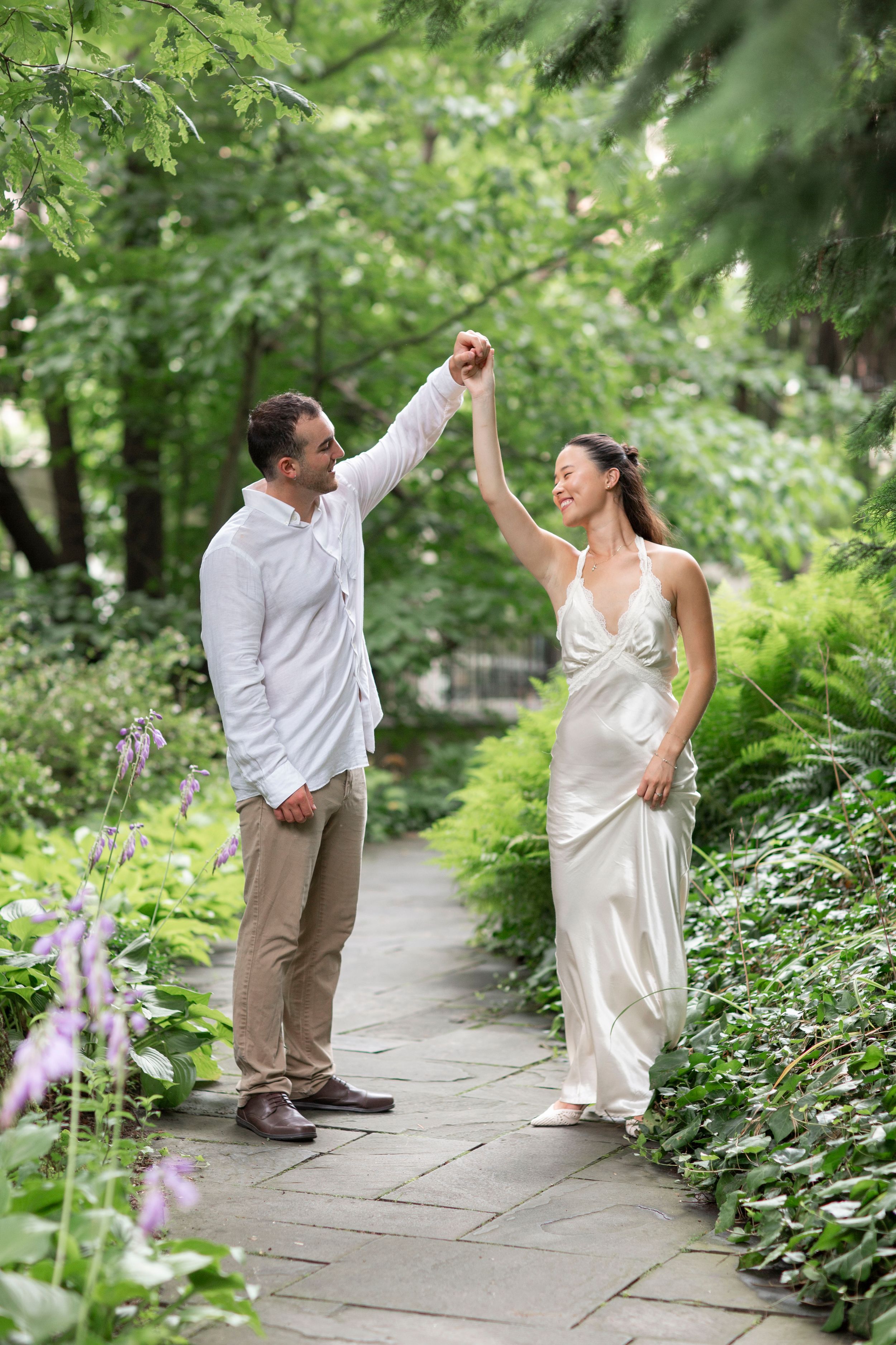 Groom twirls bride in a white satin gown along a lush garden stone pathway.