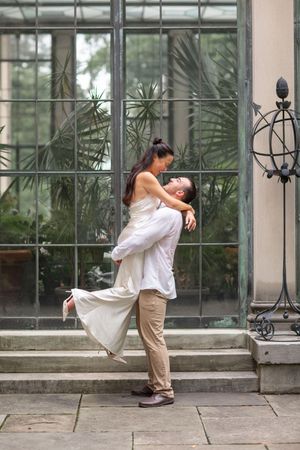 Groom joyfully lifts bride in white dress outside a glass greenhouse entrance.