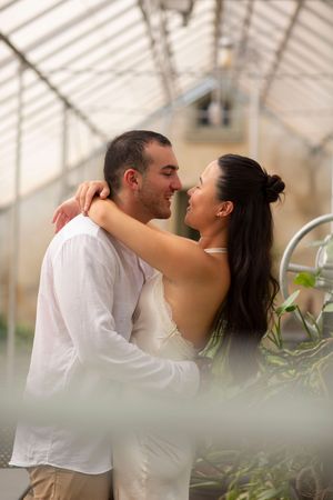 Couple shares a tender nose-to-nose moment while embracing inside a bright greenhouse.