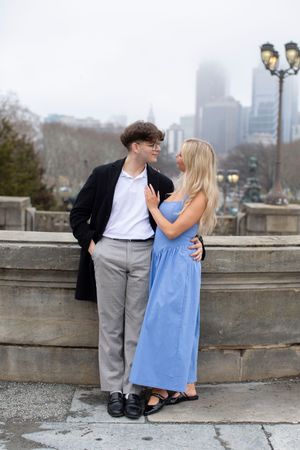 Young couple in blue dress and black coat share romantic moment at stone balustrade with city skyline.