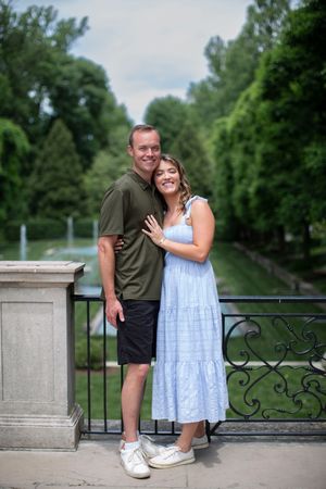 Happy couple posing on ornate bridge in lush green park, woman in blue dress showing engagement ring.