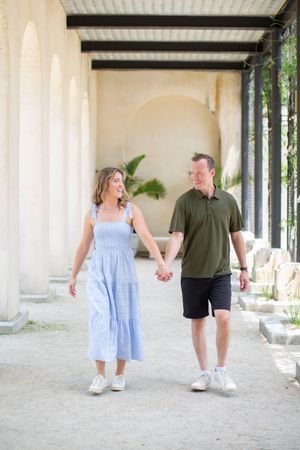 A smiling couple holds hands while walking under arched columns, woman in blue dress and man in green polo shirt.