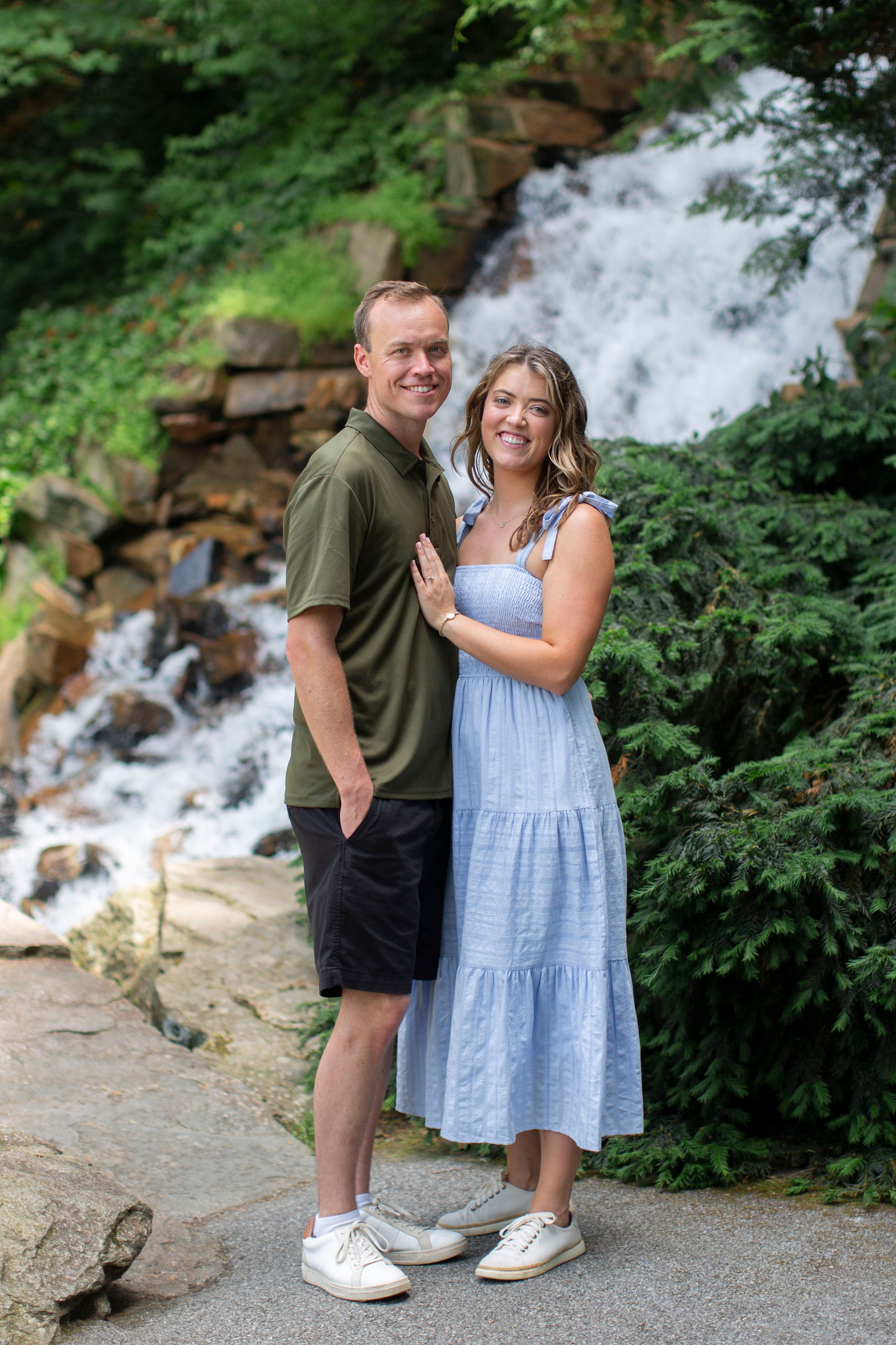 Smiling couple posing together in front of a cascading waterfall surrounded by lush green trees.