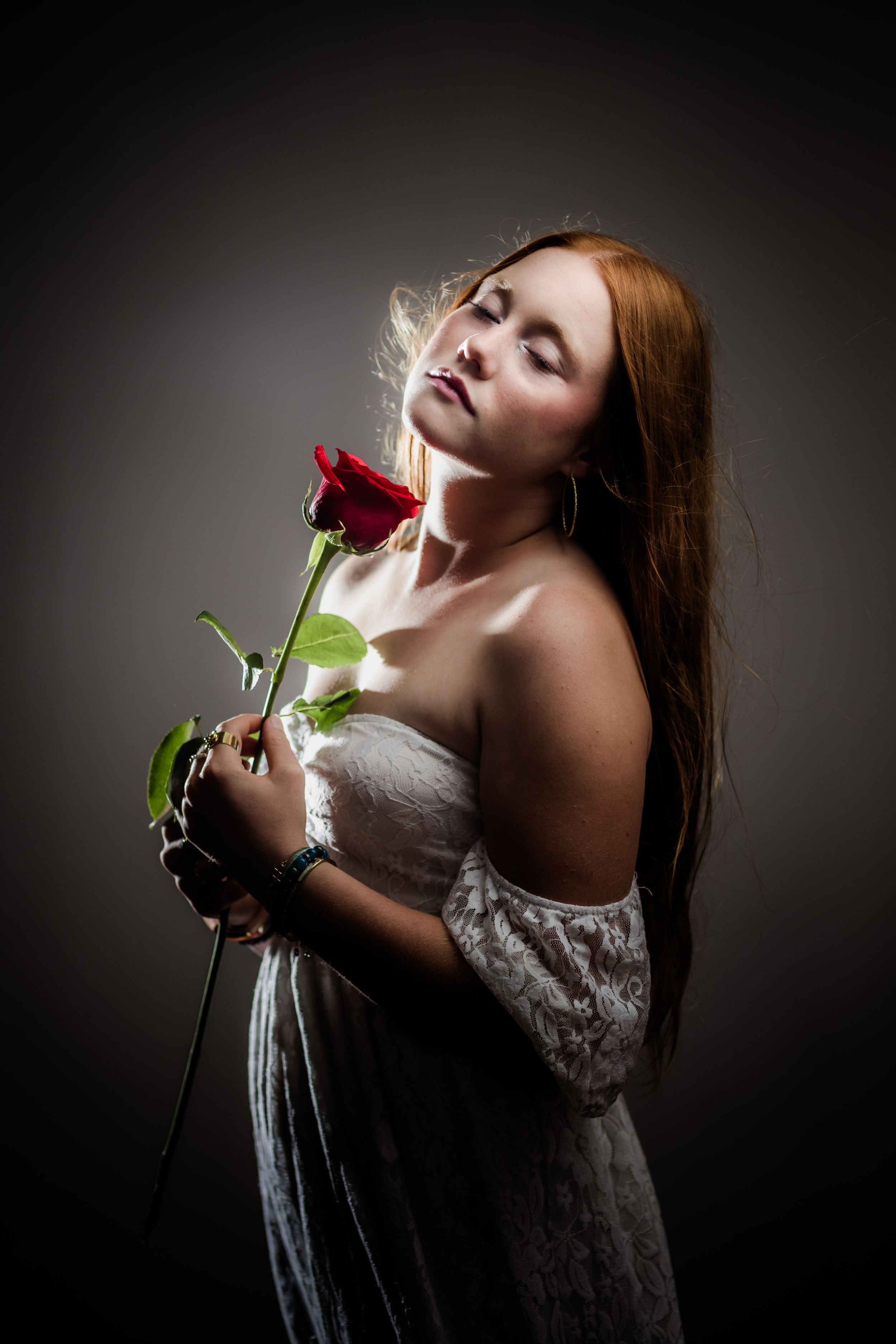 Young redhead woman in white dress holding a red rose with eyes closed against dark background.