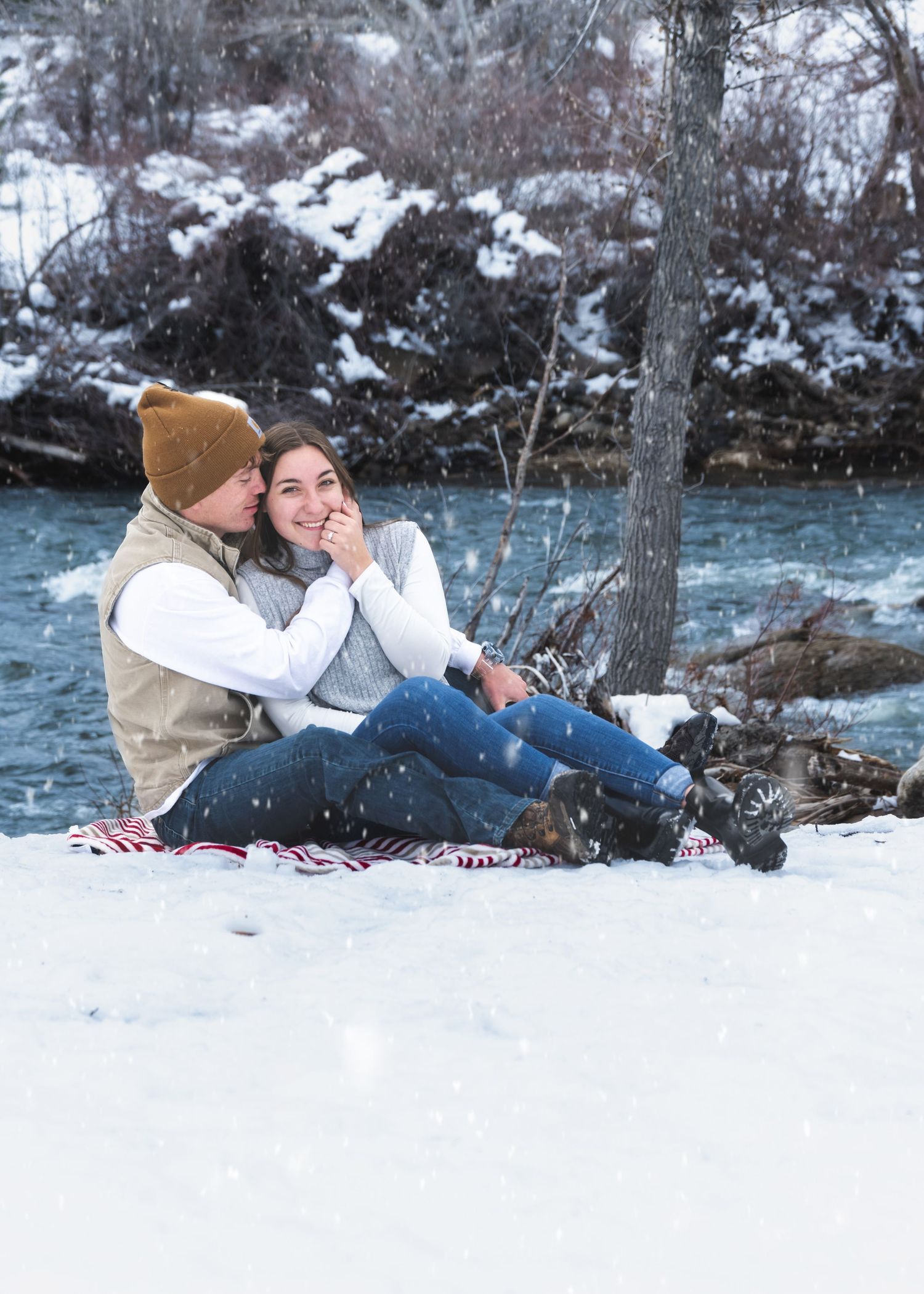Couple sitting together on snowy riverbank during winter snowfall in romantic portrait.