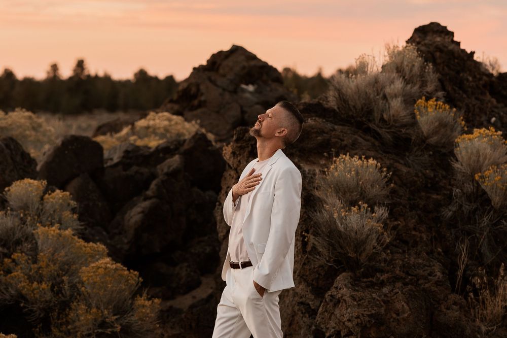 Man in white suit poses with head tilted back at sunset in rocky desert landscape.