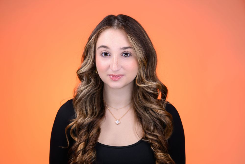 Studio portrait against orange background with someone wearing black top and wavy brown hair.