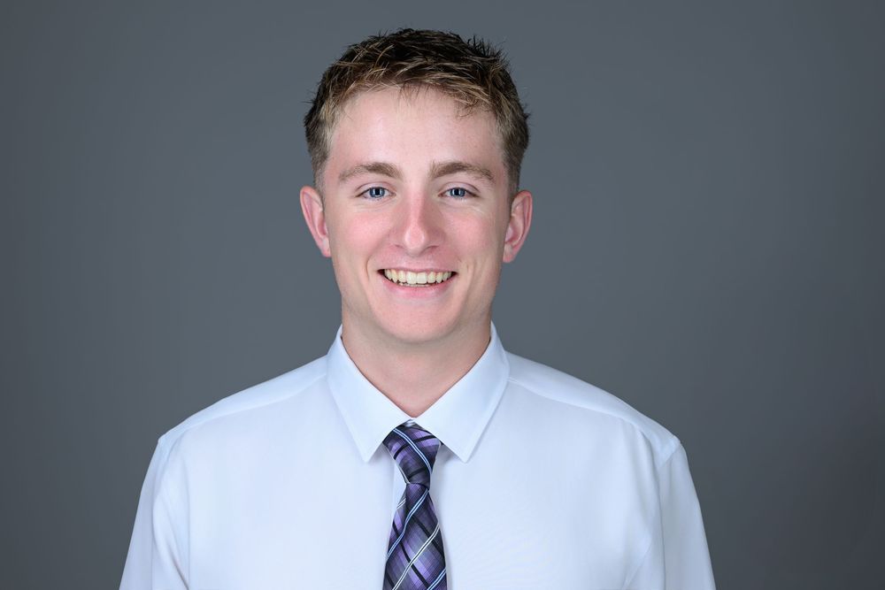 Professional headshot of a young person wearing a light blue dress shirt and purple striped tie against a gray background.