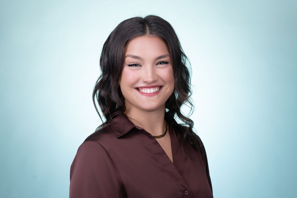 Professional headshot portrait against light blue background showing warm smile and dark wavy hair.