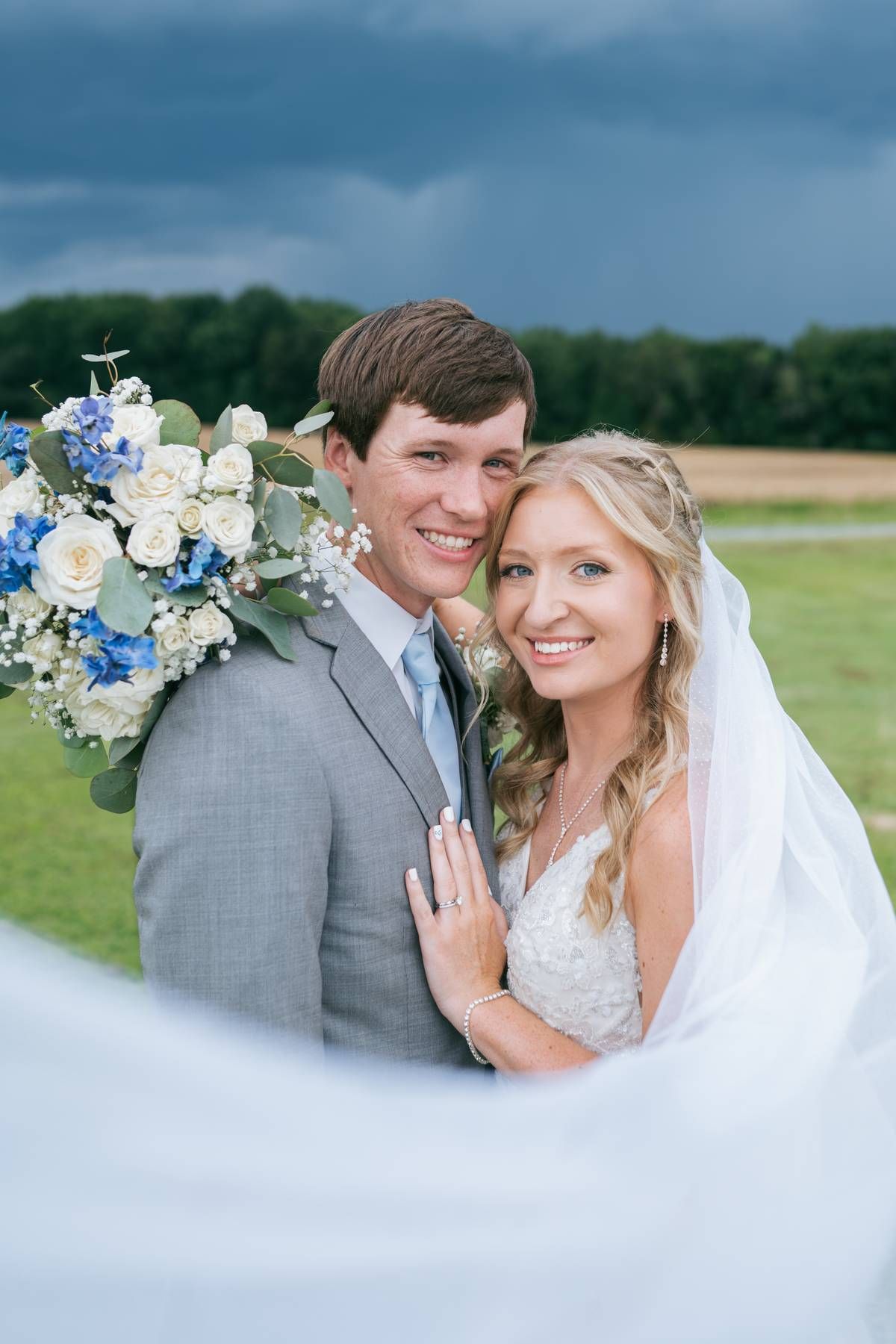 Smiling bride and groom posing with blue and white bouquet under stormy sky, wedding photographer High Point NC