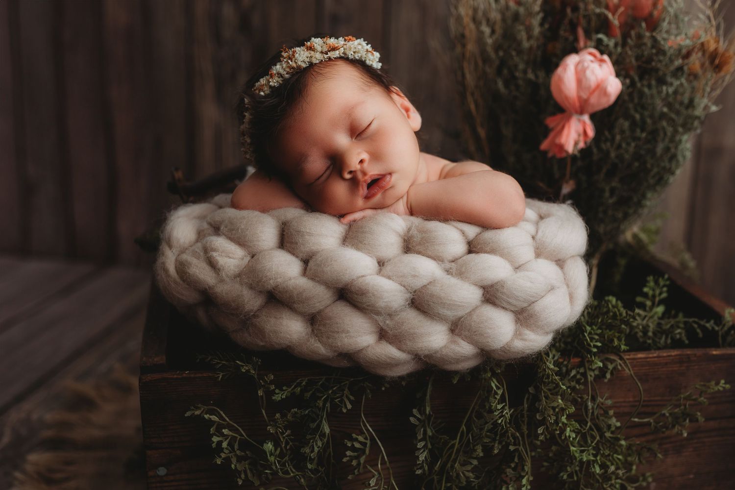 Sleeping newborn nestled in a chunky knit basket with pink flowers.