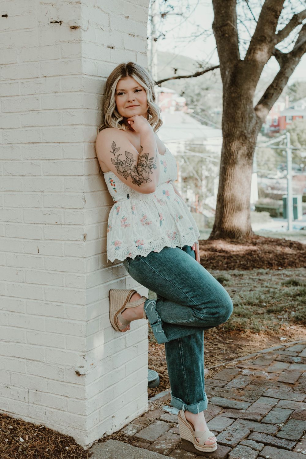 Person in floral top and jeans poses casually against white brick wall outdoors.