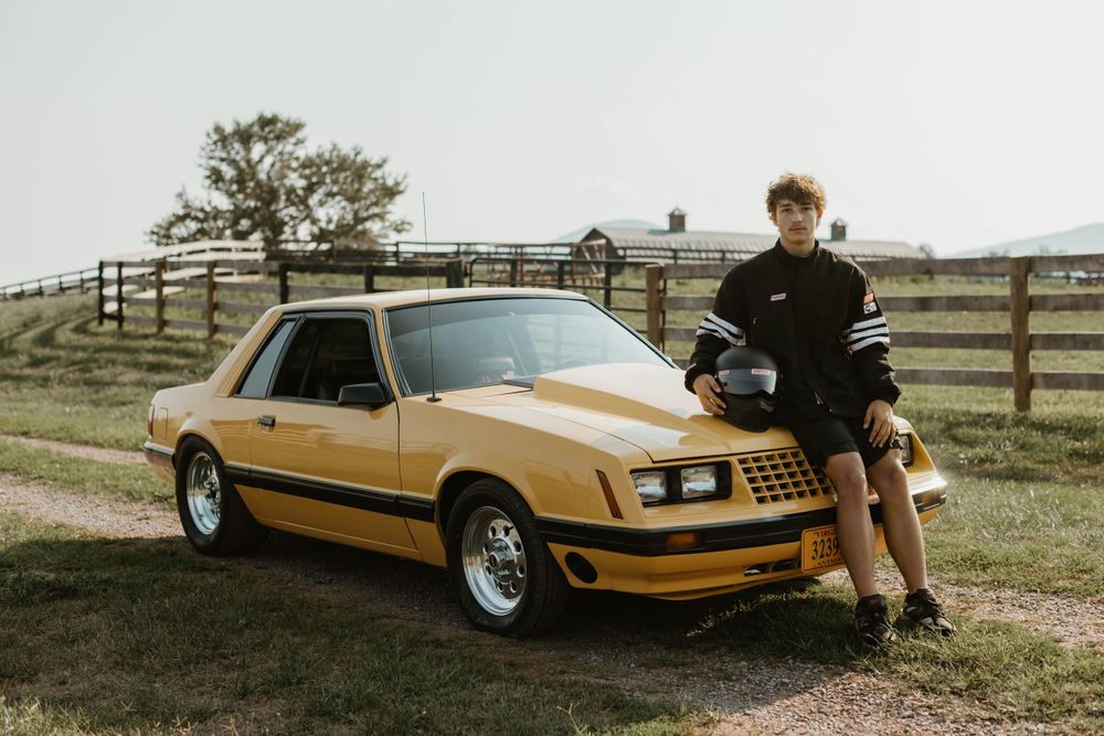 A vintage gold Ford Mustang parked in front of a wooden fence on a rural property.
